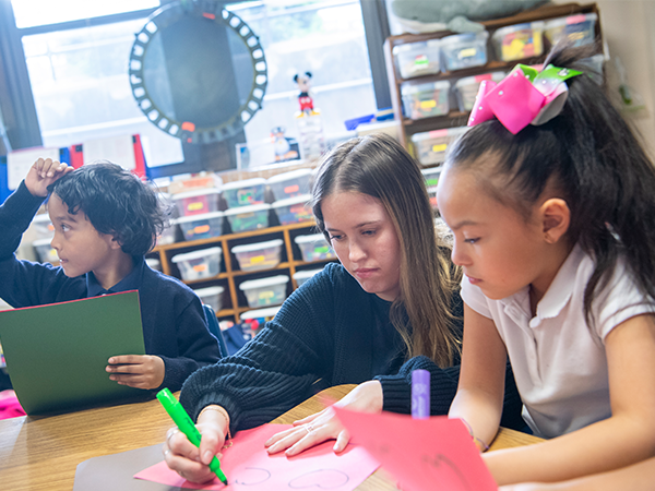 A student, kneeling down between two elementary school students, writes on construction red paper with a neon green marker in a grade school classroom.